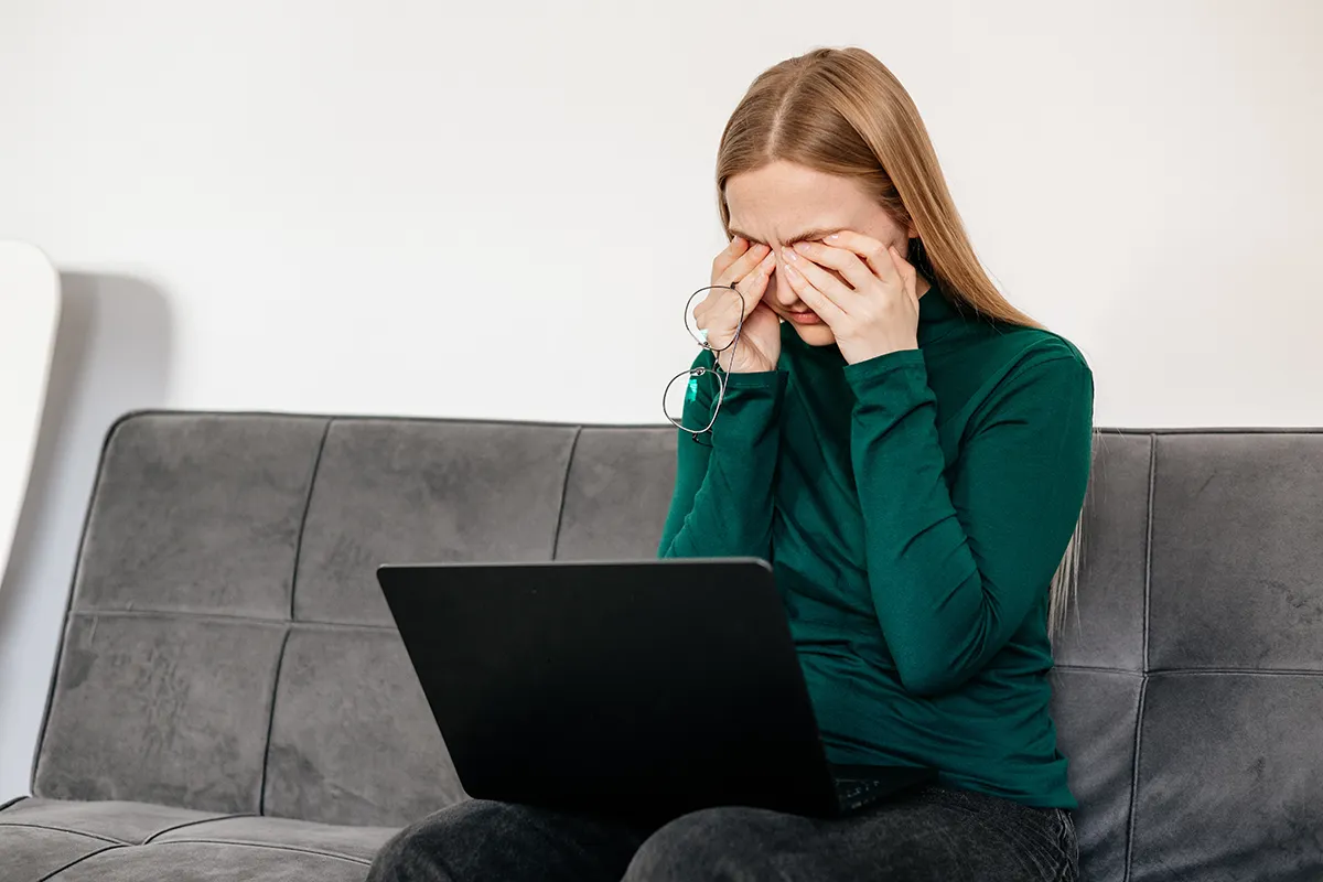 eye-strain a woman sitting on her couch with a computer on her lap and rubbing her tired eyes