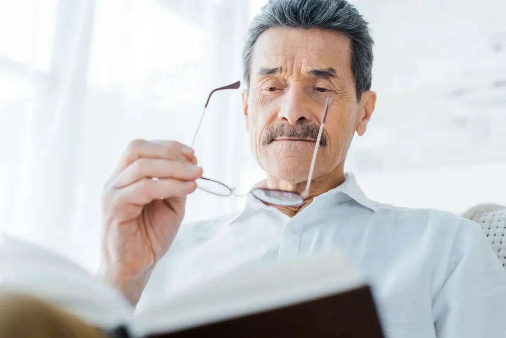 An older man reading a book. He has removed his glasses and is squinting down at the page.